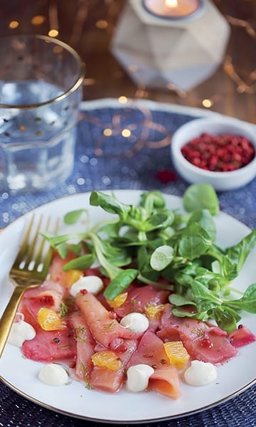 Photo prise en plongée saumon mariné au vinaigre de Reims, miel et baies roses dans une assiettes blanche, elle même dans un plat bleu. Dans ce plat on voit aussi un verra d'eau