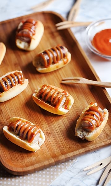Jolie photo de minis hot-dogs aux chipos et sauce barbecue express, on retrouve ce plat sur une planche en bois de dégustation, avec des petites fourchettes à escargots en bois ainsi qu'un petit pot en verre avec une petite sauce à l'intérieur