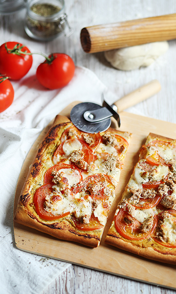Pizza sur une jolie nappe blanche et sur un plateau en bois, une roulette à pizza, un rouleau à pâtisserie, un pot de moutarde, et la pizza thon et tomate à la moutarde aux herbes de Provence